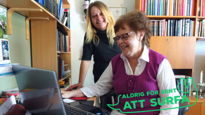 En äldre kvinna sitter framför en bärbar dator. Bakom henne står en kvinna i medelåldern som ler och ser in i kameran. På bilden finns också en logga med texten "Aldrig för sent att surfa".