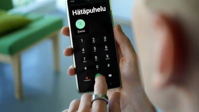 Puhelin ihmisen käsissä, näytöllä tekstit Hätäpuhelu ja Emmi sekä numeronäppäimet.