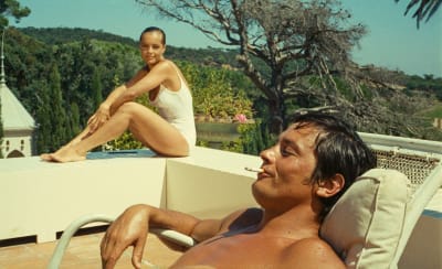 Man ligger i solstol och röker, kvinna sitter på poolkanten bakom honom