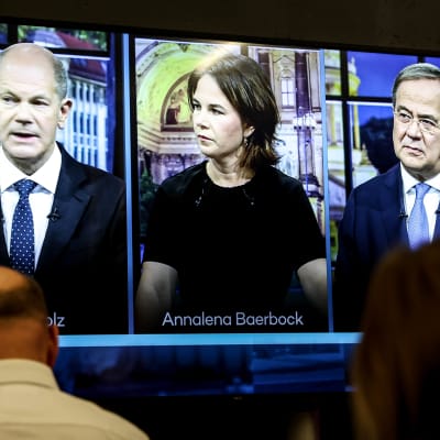 Tre tyska politiker sida vid sida på en tv-skärm. I förgrunden personer som tittar på skärmen.