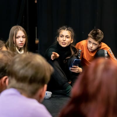 Ukrainalainen lasten- ja nuortenteatteriryhmä istuu ringissä Huoneteatterin lavalla ja leikkii teatterileikkiä. Lähikuvassa ryhmän 25-vuotias ohjaaja Maríía Ívanova Harkovasta.