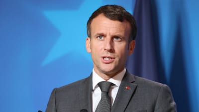 Frankrikes president Emmanuel Macron håller tal. I förgrunden syns två mikrofoner, i bakgrunden en blå bakgrundsskärm.