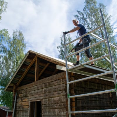 En man står på en byggnadsställning i höjd med taket på ett stockhus.