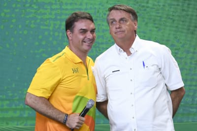 Två män står bredvid varandra: till höger Jair Bolsonaro i kortärmad vit skjorta, till vänster sonen Flávio i kortärmad gul skjorta.