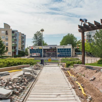 En avstängd park med trappor framför Mellugnsbacka metrostation. Parkens stenbeläggning är inte i skick, och det står högar av kakel i parken. En pojke klättrar på en träkonstruktion i parken.