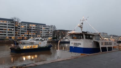 En blåvit sjöbuss, M/S Hamnskär, åker förbi en annan sjöbuss, M/S Jaarli, i Aura å i Åbo en smådimmig morgon.