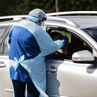 En person i skyddsutrustning tar ett coronatest på en person i en bil.