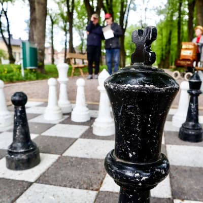 Stora schackpjäser i närbild. Två män står och ser fundersamt på schackbrädet.