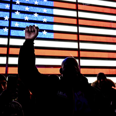 Etualalla miehiä, joista yksi nostaa nyrkkiään pystyyn, ja tautalla Yhdysvaltain lippu näytöllä.