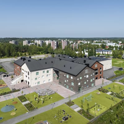 Harjakattoinen rakennus, jossa on punaisia, valkoisia ja harmaita osia. Niiden etualalla on vaaleanvihreää ruohoa neliskanttisina alueina, joilla on myös leikkivälineitä. Taustalla näkyy metsää ja kerrostaloja. 