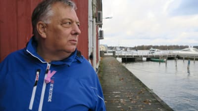 En man med grått hår och blå jacka (Mats Lagerstam) står på en brygga vid båthamnen i Ekenäs. Stallörsparken. Bakom ser man lite av ett rött trähus.
