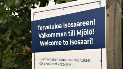 En vit skylt med texten "Välkommen till Mjölö" på blå bakgrund, skrivet på finska, svenska och engelska. Bakom sklyten syns gröna löv.