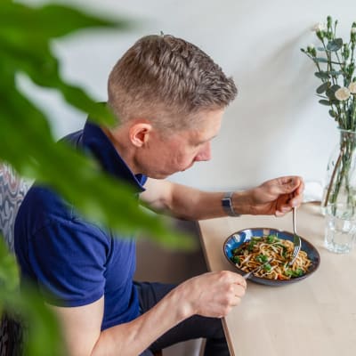Klaus Raunela istuu pöydässä ja edessään hänellä on kasvisruokaa lautasella. Etualalla viherkasvinlehtiä.