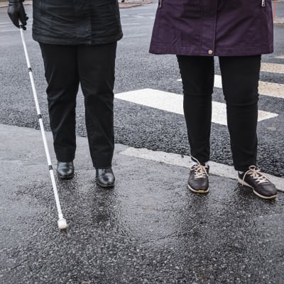 Benen av två personer som står intill ett övergångsställe. Den ena person har i handen en vit käpp som används för personer med synnedsättning.