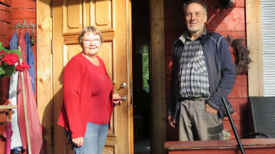 En äldre kvinna och äldre man står framför en rödmålad stockstuga. De ser in i kameran och kvinnan öppnar dörren till huset.