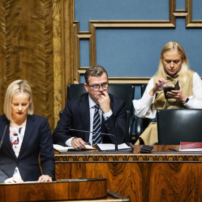 Riikka Purra står vid ett talarpodium och pratar. I bakgrunden sitter Petteri Orpo och Elina Valtonen.
