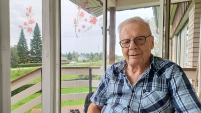 En gammal man i rutig skjorta sitter på en glasveranda (Rune Björkqvist)