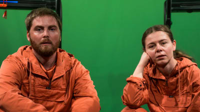 Skådespelarna Ole Øwre och Elisa Makarevitch sitter på marken iklädda orange overaller, i bakgrunden syns en grön skärm.