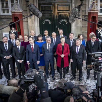 Danmarks nya regering utanför Amalienborg slott i Köpenhamn med statsminister Mette Frederiksen i mitten. Gruppen på 23 personer är omgiven av kameror och mikrofoner och bakom dem står två soldater på ömse sidor om en dörr.