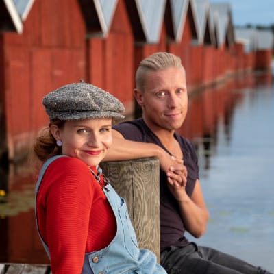 Programledarna Hannamari Hoikkala och Nicke Aldén sitter på en brygga vid en sjö, en rad med rödmålade fiskebodar i bakgrunden.