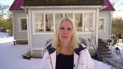 Jessica Lindgren framför sitt hus i soligt vinterväder. Hon har blont långt hår och en vit jacka.