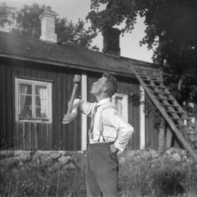 En man leker leken "kyssa gudfar", där han lyfter en slägga med en arm och kysser släggan. Bilden är tagen 1930 i Bromarf.