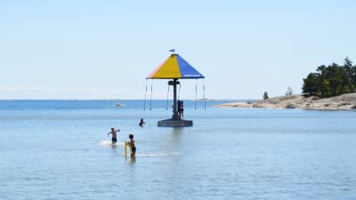 En bild på havet. I mitten av bilden finns en karusell som snurrar i vattnet. barn åker i karusellen och barn springer i grunt vatten. Strandidyll.