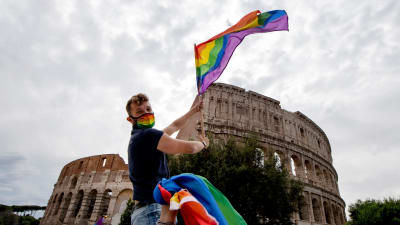 En man sitter på ett biltak och håller i en prideflagga. I bakgrunden romska colosseum. 