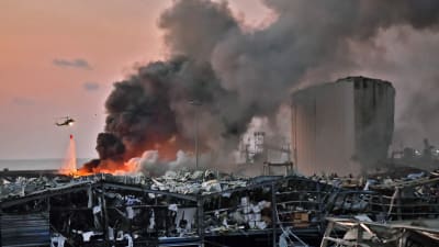 Vyn visar förstörda byggnader brand i hamnen efter explosionen i Beirut. Rök stiger upp från huset och en helikopter som deltar i släckningsarbetena flyger förbi.
