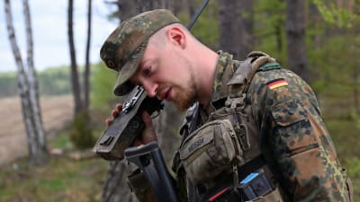 En tysk soldat i kamouflagedräkt och keps använder en radiotelefon i en skog under en övning i Polen den 6 maj 2022.. 