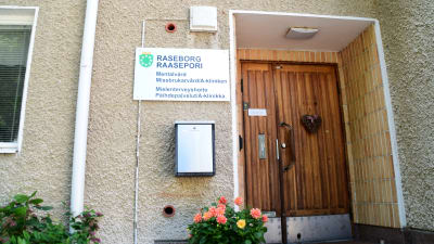 En bild på en dörr på ett hus. En blomma står på trappan och ett hjärta hänger på dörren. På en skylt står det Raseborgs Mentalvård, Missbrukarvård/A-kliniken på finska och svenska.