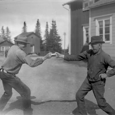 Två män leker leken "dra dosa", där båda håller i en och samma snusdosa mellan sina pek- och långfingrar och försöker dra den åt sig själv. Bilden är tagen 1931 i Närpes, Näsby.