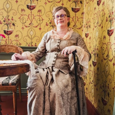 Piia Vähäsalo sitter på handelshusets cafésida iklädd en klänning i äkta 1800-tals anda samtidigt som hon lutar sig mot sin gångkäpp.