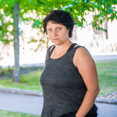 En man och en kvinna står och tittar allvarsamt in i kameran. I bakgrunden syns träd, många gröna löv.