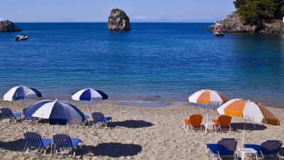 Bild på strand med blåvita och orangevita parasoll och solstolar.
