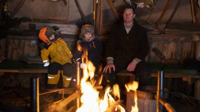 En pappa och två små pojkar sitter vid en eldstad.