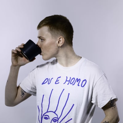 Edith Hammar står i en vit studio och dricker ur en kaffemugg, hon har en vit t-skjorta som det står "Du e homo" på.