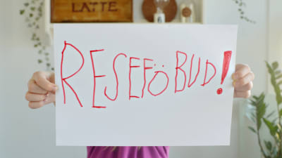 Ett barn håller upp ett stort pappersark med texten "RESEFÖBUD!".