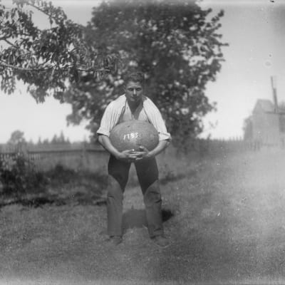 En man lyfter en stor sten med sina händer. Bilden är tagen 1930 i Jomala, Björsby, Åland.