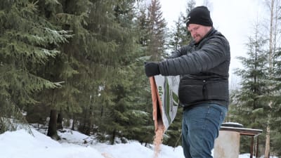 En jägare i svart jacka och svart luva häller ut ärter ur en stor påse i en snöig skog för att mata vitsvanshjortar med.
