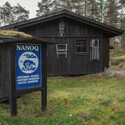 Sinivalkoinen kyltti jossa teksti Nanoq ja jääkarhun kuva, tummia mökkejä taustalla