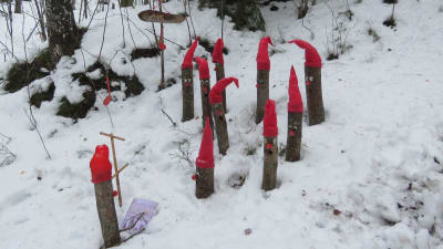 "Jultomtar" gjorda av stående trästockar iklädda röda tomteluvor och dekorerade med ögon och en röd näsa. Föreställer en tomtekör som leds av en dirigent.