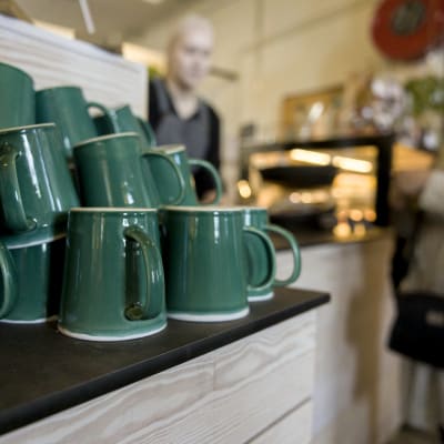 Gröna kaffekoppar staplade på varandra, i bakgrunden skymtar en cafédisk med en anställd och en kund i samspråk.