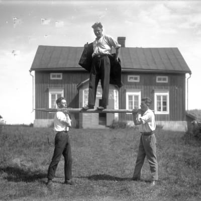 En man balanserar på en stång som bärs av två män. Bilden är tagen 1930 i Jomala, Åland.