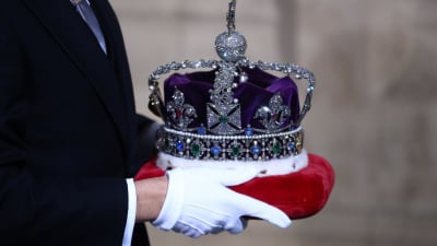 Händer som håller i Drottningens Imperiestatskrona på en röd dyna.