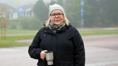 En kvinna med svart rock och grå mössa och en termosmugg i handen står utomhus, en dimmig morgon. Sjundeå station/centrum. Hon håller också en tidning i sin andra hand.