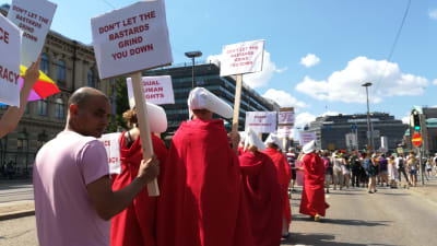 Människor på demonstration inför toppmötet mellan Trump och Putin som är klädda i röda kappor och vita huvudbonader som påminner om tv-serien Handmaid's tale.