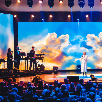 Katri Helena laulaa Kulttuuritalon lavalla täydelle yleisölle. Helenalla on päällään valkoinen asu. Lavalla näkyy myös neljä muuta muusikkoa. Taustalla on kuva taivaasta, jossa on pilviä ja auringonsäteitä.