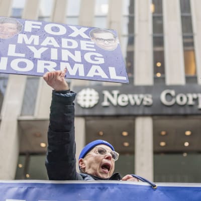 En person håller upp en skylt där det står "Fox made lying normal" utanför en byggnad.
