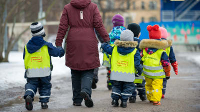 Dagisbarn och personal på promenad 25.3.2021. Man ser bara ryggarna av dem. Barnen har neongula reflexvästar på sig.